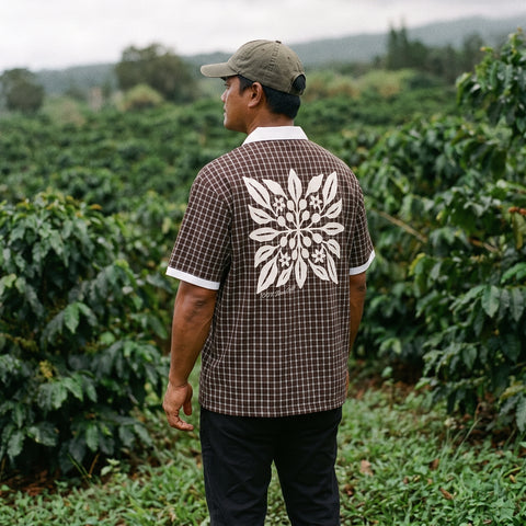 A man standing in a lush green outdoor field, viewed from behind, wearing a brown and white checkered short-sleeve shirt with a large white coffee floral pattern on the back, black pants, and a khaki cap.