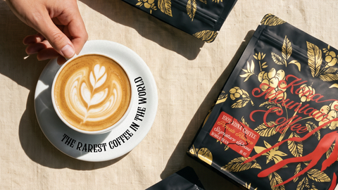 A hand holds a white cup of latte art coffee on a saucer beside branded coffee pouches on a beige fabric surface. The packaging is black with gold floral illustrations and red text, and the cup reads 'THE RAREST COFFEE IN THE WORLD.