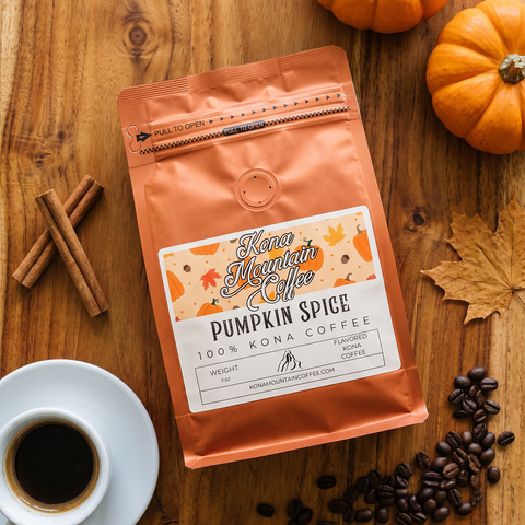 A bag of Kona Mountain Coffee Pumpkin Spice coffee lies on a wooden tabletop, surrounded by a cup of black coffee, cinnamon sticks, coffee beans, autumn leaves, and small pumpkins. The bag is metallic orange with a white and orange label.